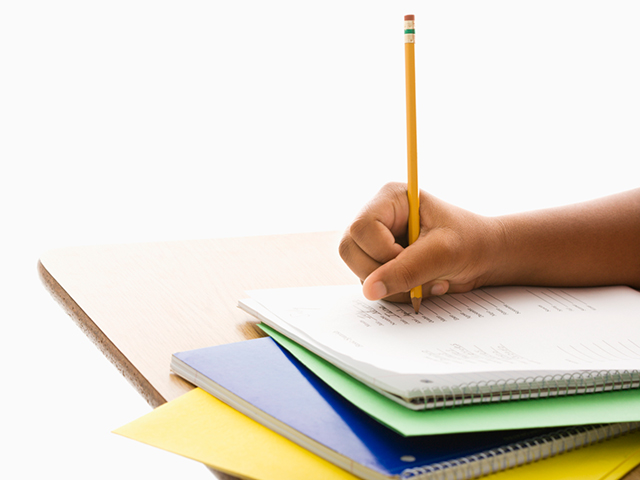 Close-up analog photograph of a hand holding a pencil writing in a notebook on a wooden desk with stacked notebooks